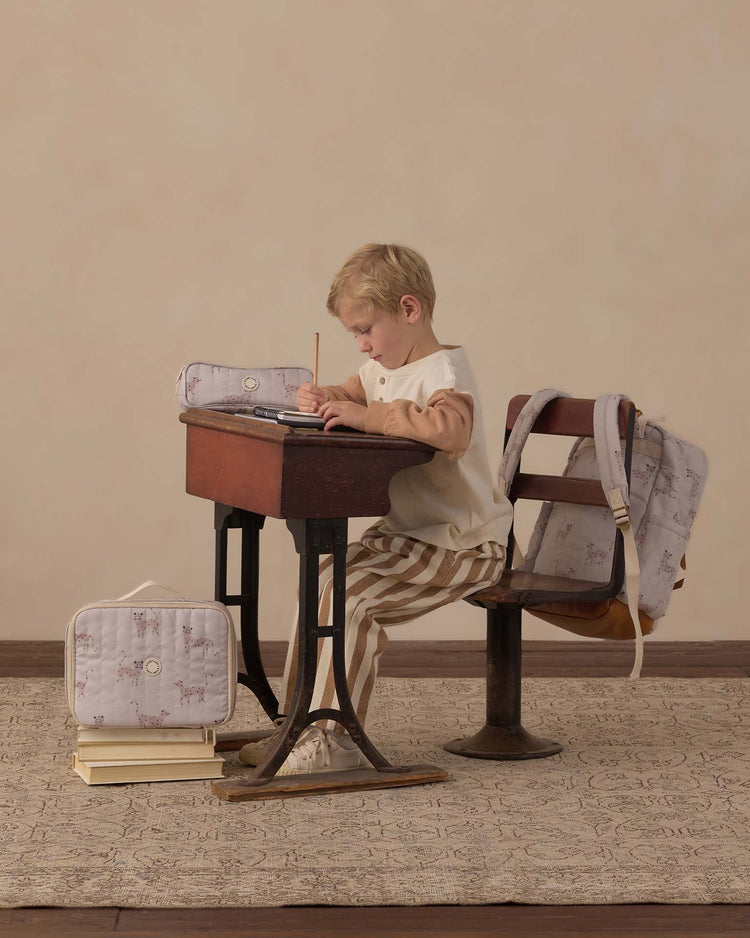 A young child sits at a vintage school desk, drawing with a pencil. Nearby are a backpack, lunchbox, and the rylee + cru pencil pouch in leopard—classic school essentials with matching patterns. The child wears striped pants and a sweater, creating a warm, nostalgic atmosphere.