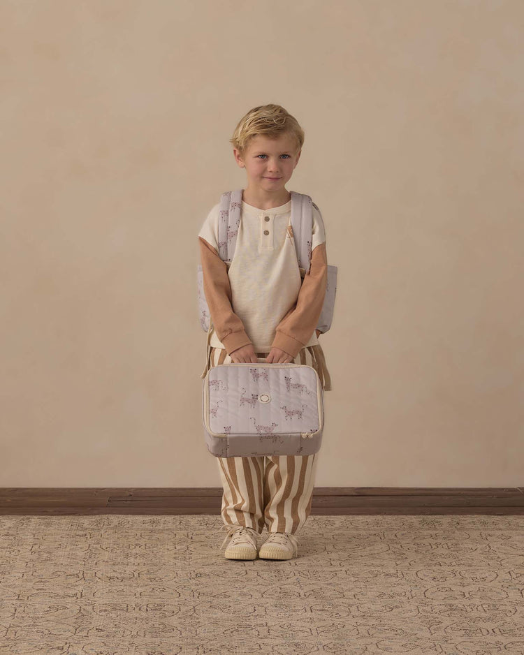A young child with blonde hair stands indoors against a plain beige background, dressed in a cream shirt and brown striped pants, holding a matching animal-print backpack and the rylee + cru lunch bag in leopard.
