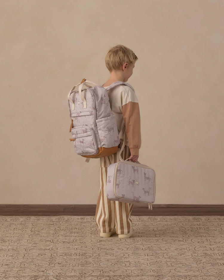 A young child stands sideways, ready for everyday adventures, wearing the rylee + cru backpack in leopard—crafted from durable polyester—and holding a matching lunchbox. The child wears a brown and cream sweater with striped pants, standing on a textured carpet against a plain beige wall.