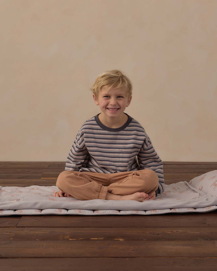 A smiling young child with blond hair sits cross-legged on the rylee + cru nap mat in leopard, featuring a removable pillow and soft micro-fleece blanket, on a wooden floor. The child wears a striped long-sleeve shirt and peach pants, pictured against a plain beige background.