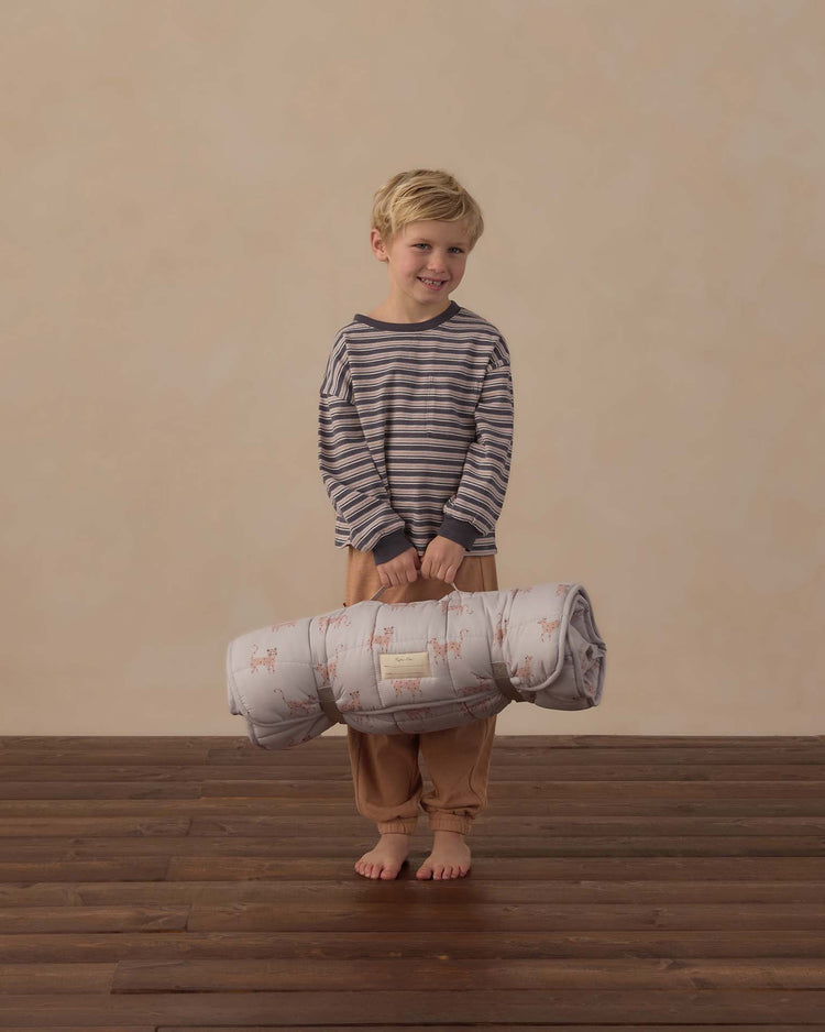 A young child in striped pajamas stands barefoot on a wooden floor, holding a rylee + cru nap mat in leopard with both hands—the micro-fleece blanket peeking out—smiling at the camera against a plain beige background.