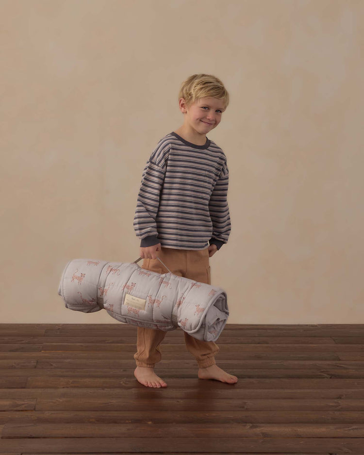 A young boy in a striped shirt and tan pants stands barefoot on a wooden floor, smiling and holding the rylee + cru nap mat in leopard by its handle. The background is plain and beige.