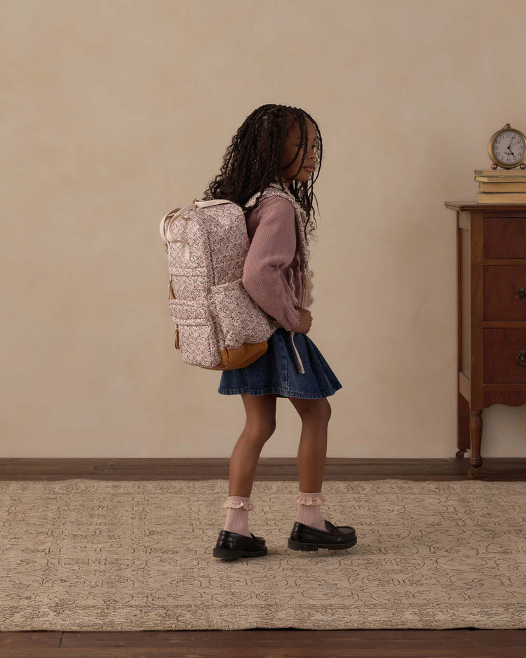 A young girl with long braids wears a pink sweater, denim skirt, and the rylee + cru backpack in primrose with its spacious main compartment, standing on a rug beside a wooden dresser topped with books and a clock—ready for everyday adventures.