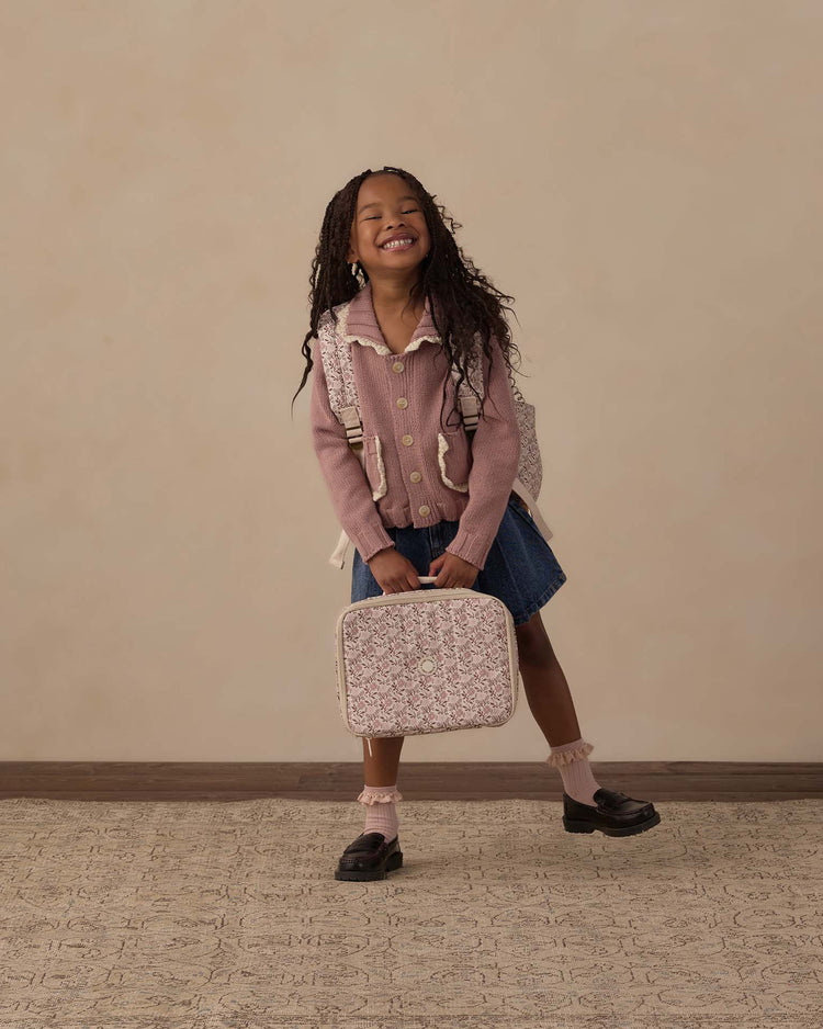 A smiling young girl with long braids, dressed in a pink sweater, denim skirt, and black shoes, stands holding the rylee + cru lunch bag in primrose and wears a matching backpack, posing against a neutral background.