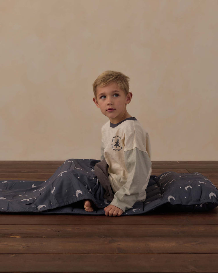 A young boy with blonde hair sits on a wooden floor, comfortably nestled on his rylee + cru nap mat in moons. Wrapped in a soft, dark gray micro-fleece blanket adorned with white patterns, he wears a beige long-sleeve shirt and looks slightly to the side with a neutral expression.