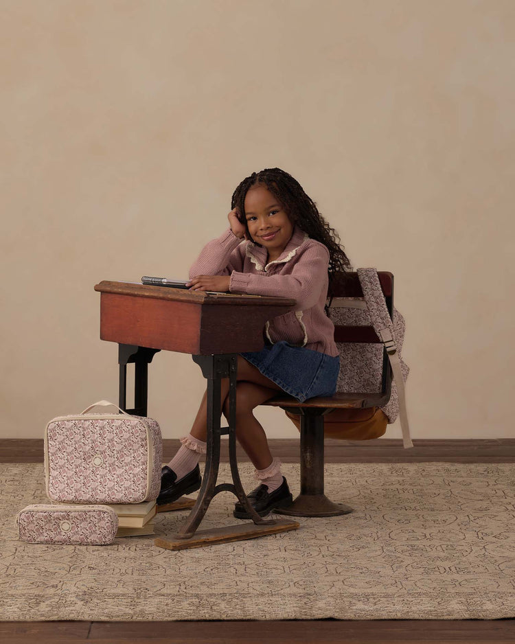A smiling young girl with long hair sits at an old-fashioned wooden school desk, wearing a pink sweater and denim skirt. A rylee + cru pencil pouch in primrose lies among patterned bags filled with school essentials on the floor, while a backpack hangs from the chair. The scene is set against a neutral background atop a patterned rug.