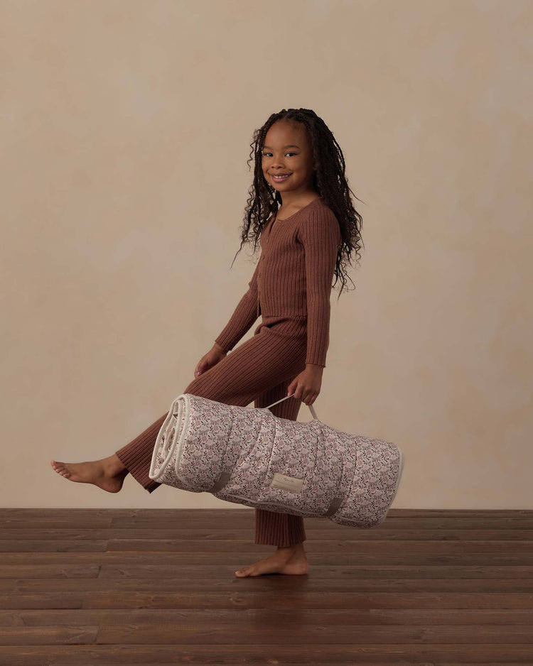 A smiling child in brown pajamas holds the rylee + cru nap mat in primrose, standing barefoot on a wooden floor against a beige background.