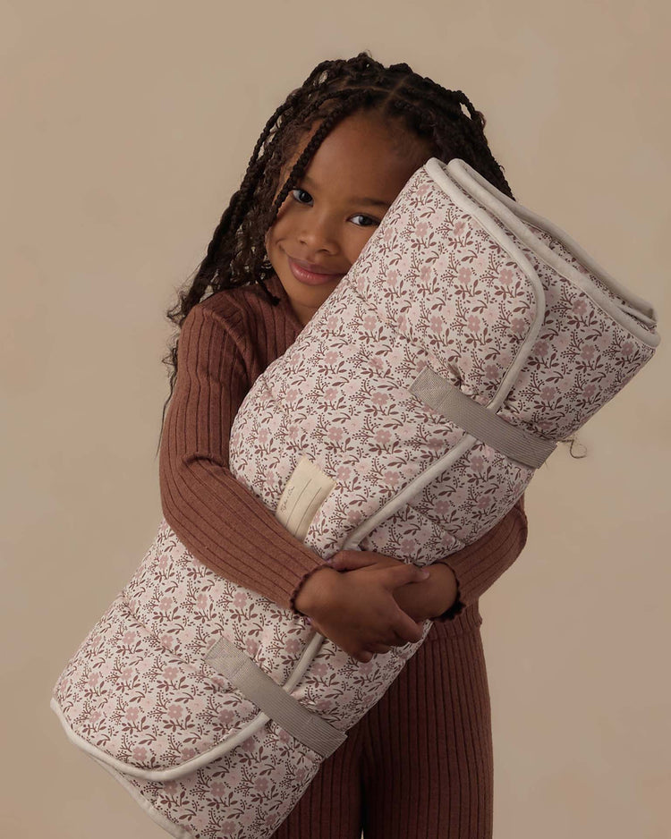 A young child with long braids smiles while hugging the rylee + cru nap mat in primrose, which features a soft micro-fleece blanket for cozy comfort. The child wears a ribbed brown outfit against a plain beige background.