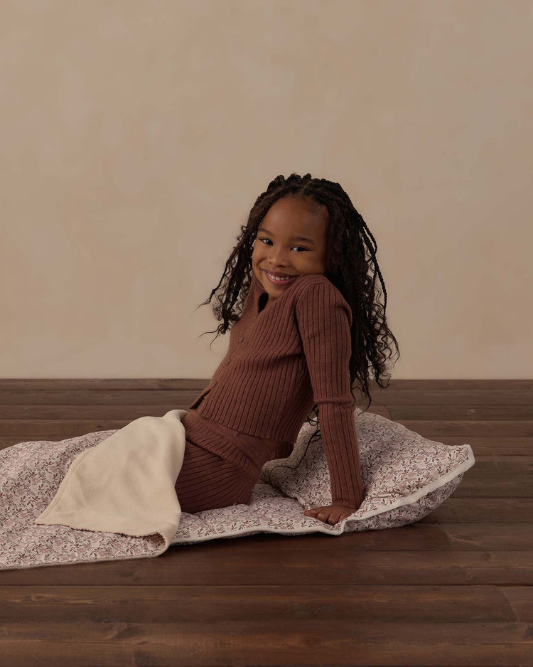 A young girl with long braided hair sits smiling on a wooden floor, wearing a brown ribbed outfit. She is seated on the rylee + cru nap mat in primrose, which features a cream-colored section. The background is plain beige.