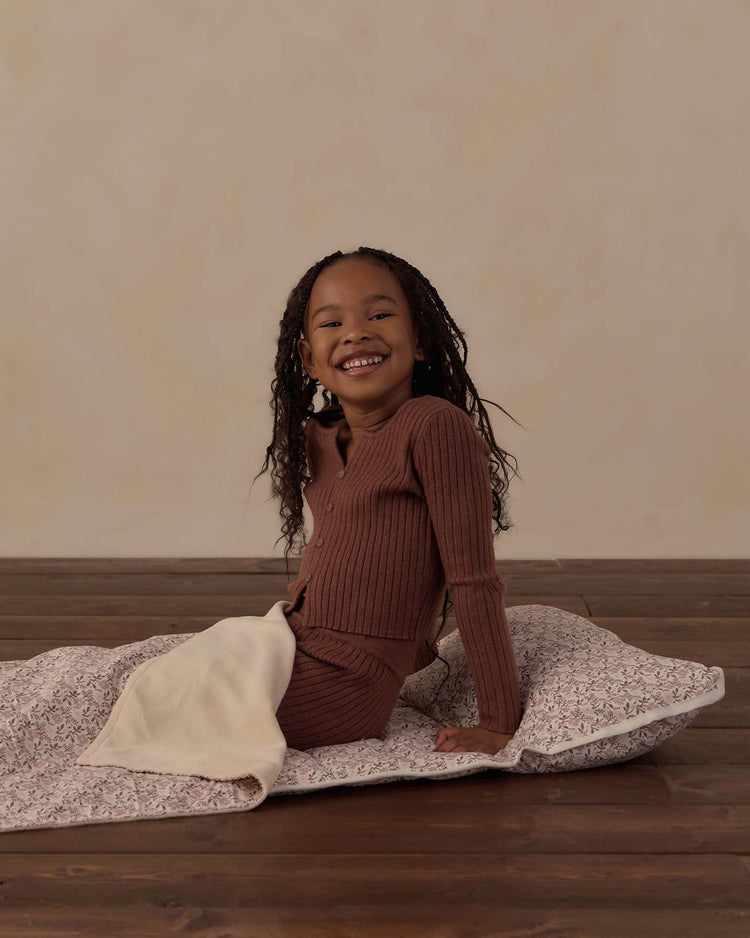 A smiling child with long hair sits on a wooden floor, wearing a brown ribbed outfit, atop the rylee + cru nap mat in primrose. The patterned nap mat features a cozy micro-fleece blanket and a removable pillow, set against a plain, light-colored wall.