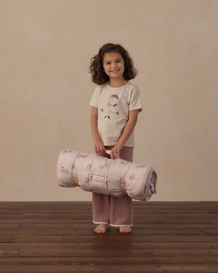 A young girl with curly hair smiles while holding a rolled-up rylee + cru nap mat in owls. She is wearing owl-print pajamas, standing on a wooden floor against a plain beige background.