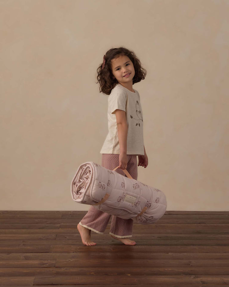 A young girl in pajamas stands on a wooden floor, smiling and holding the rylee + cru nap mat in owls by its handle. The background is plain and beige.
