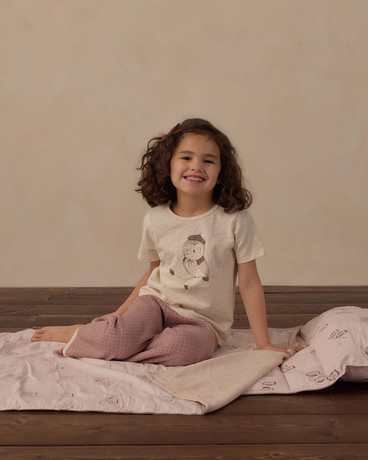 A smiling young girl with curly hair sits on a rylee + cru nap mat in owls, which features a cozy micro-fleece blanket and removable pillow, on a wooden floor; she’s wearing a beige animal-print t-shirt and pink pajama pants.