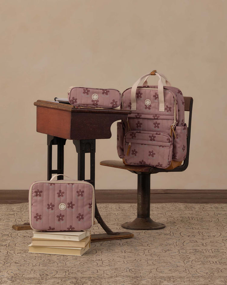 A vintage wooden school desk features a matching pink floral backpack, a rylee + cru lunch bag in little daisy, and a coordinating pencil case on and around it, with stacked books resting on the floor—all set against a plain beige wall.