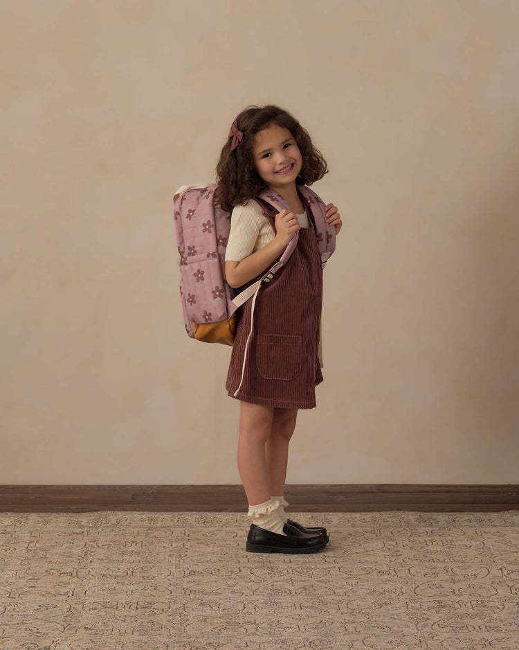 A young girl with curly hair stands smiling against a beige background, wearing a brown pinafore dress, cream shirt, and black shoes. She carries the rylee + cru backpack in little daisy—a durable polyester backpack with floral designs that's perfect for everyday adventures.