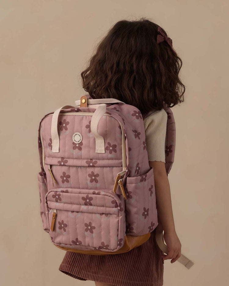 A young girl with wavy brown hair, dressed in a cream shirt and mauve skirt, carries the rylee + cru backpack in little daisy—crafted from durable polyester and adorned with charming brown flower patterns—making it perfect for everyday adventures. She is pictured facing away from the camera against a beige background.