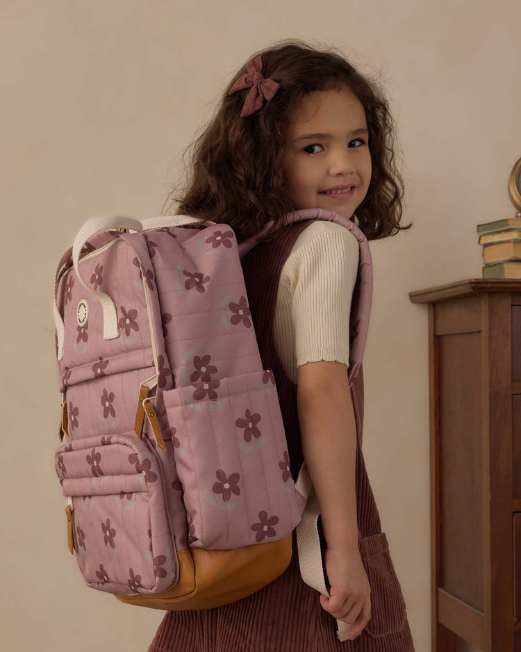 A young girl with curly hair, dressed in a cream shirt and brown pinafore, smiles as she carries the rylee + cru "backpack in little daisy"—a durable polyester pink backpack featuring charming brown flower prints—standing near a wooden dresser with a stack of books, always ready for her everyday adventures.