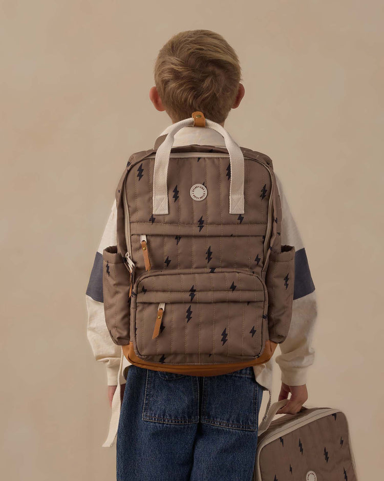 A child with short hair, dressed in a beige and navy sweater and blue jeans, stands facing away. They carry the rylee + cru "backpack in bolts"—a brown backpack featuring a lightning bolt pattern made from durable polyester—and hold the matching lunchbox in their right hand, ready for everyday adventures.