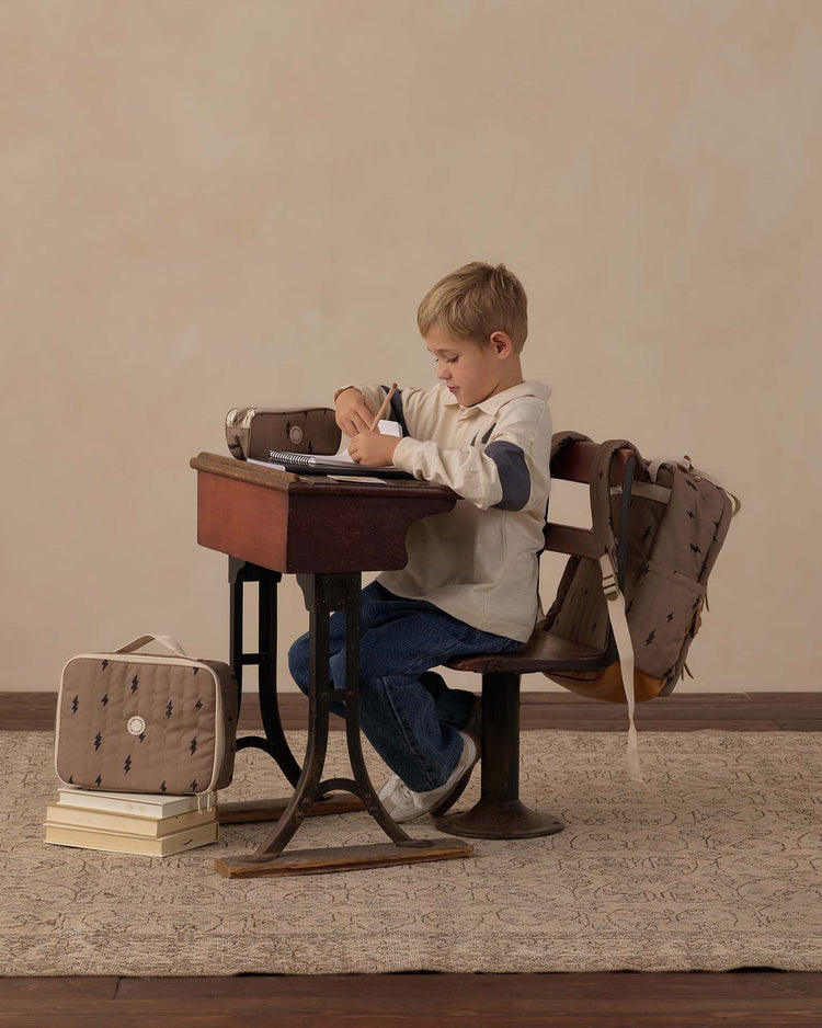 A young boy sits at a wooden desk writing in a notebook. A backpack hangs on his chair, and the rylee + cru "pencil pouch in bolts," filled with school essentials, rests atop stacked books nearby. The background is plain beige.