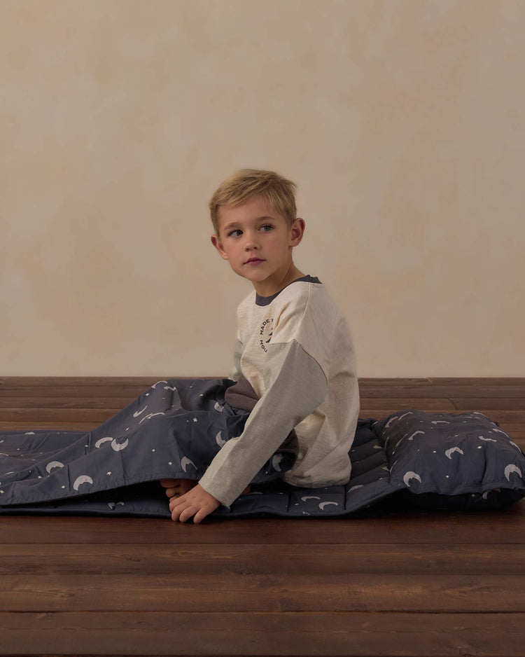 A young boy sits on the rylee + cru nap mat in moons, which features a dark blue design with white moon patterns, a soft micro-fleece blanket, and a removable pillow. He is indoors on a wooden floor with a plain beige background, looking to his left.