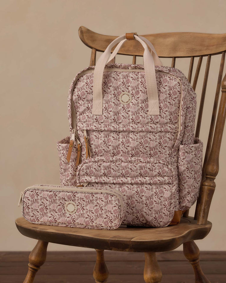 A pencil pouch in primrose by rylee + cru, decorated with pink floral patterns, sits atop a wooden chair against a beige background, making it a perfect school essential.