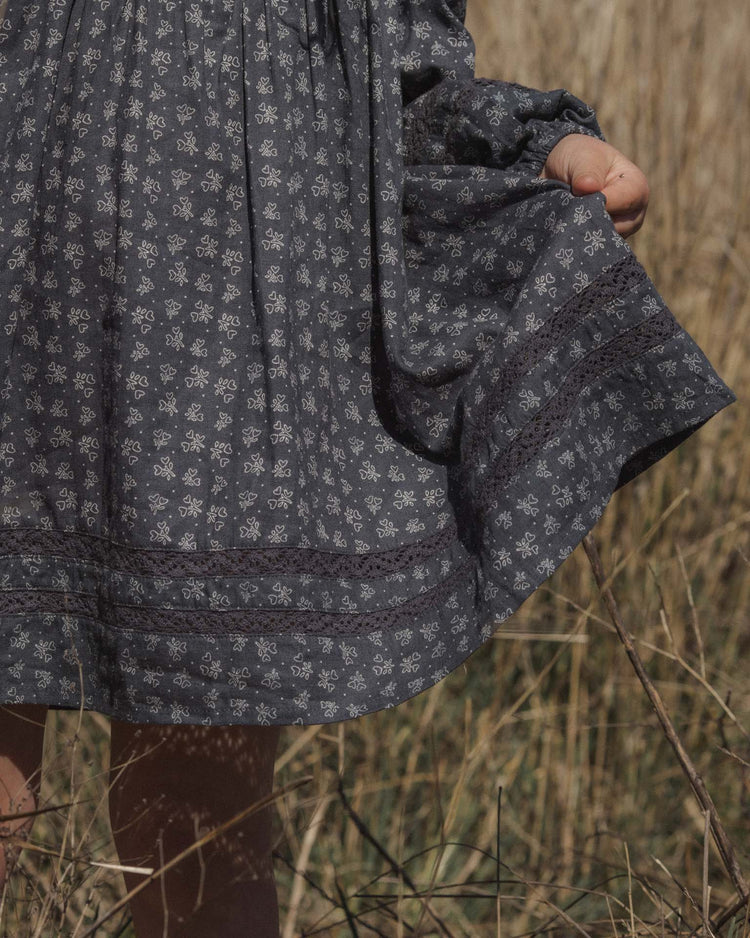 A person wearing the rylee + cru selene dress in heart ditsy stands in a dry field, holding out the hem with one hand. Only part of the patterned gray dress, along with their arm and legs, is visible.