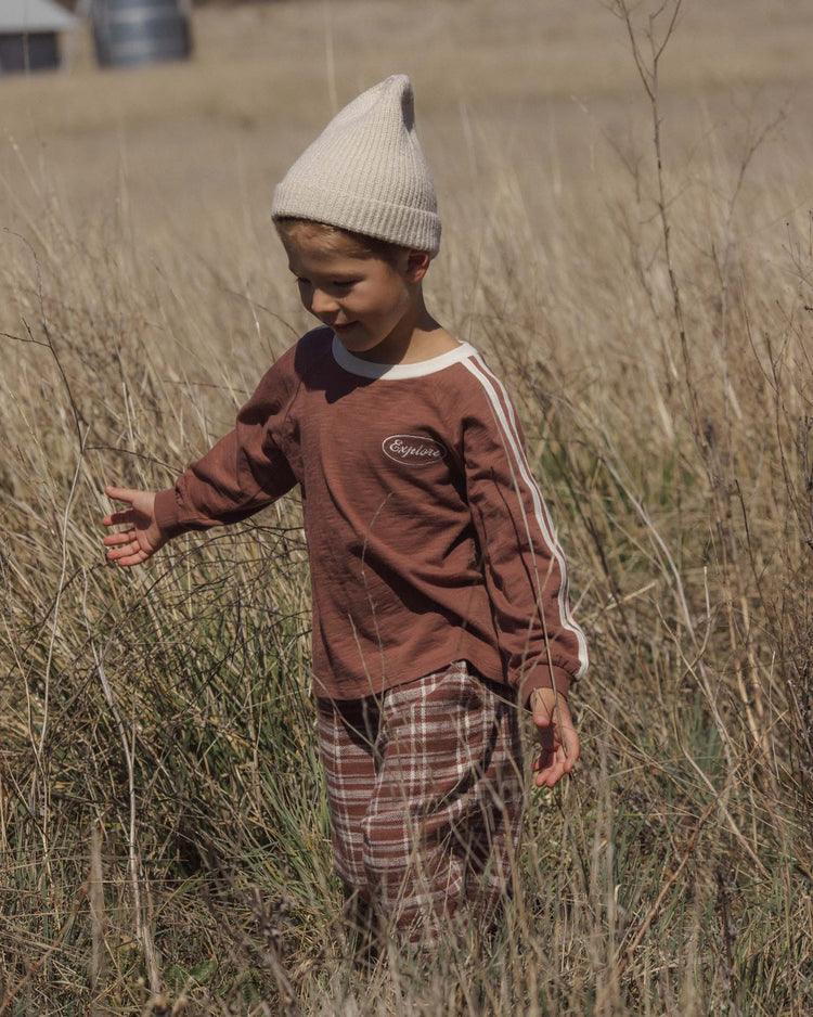 A child wearing a cream knit hat, a brown long-sleeve shirt, and the rylee + cru palmer pant in aspen plaid with an elastic waistband walks through tall, dry grass in a field on a sunny day.