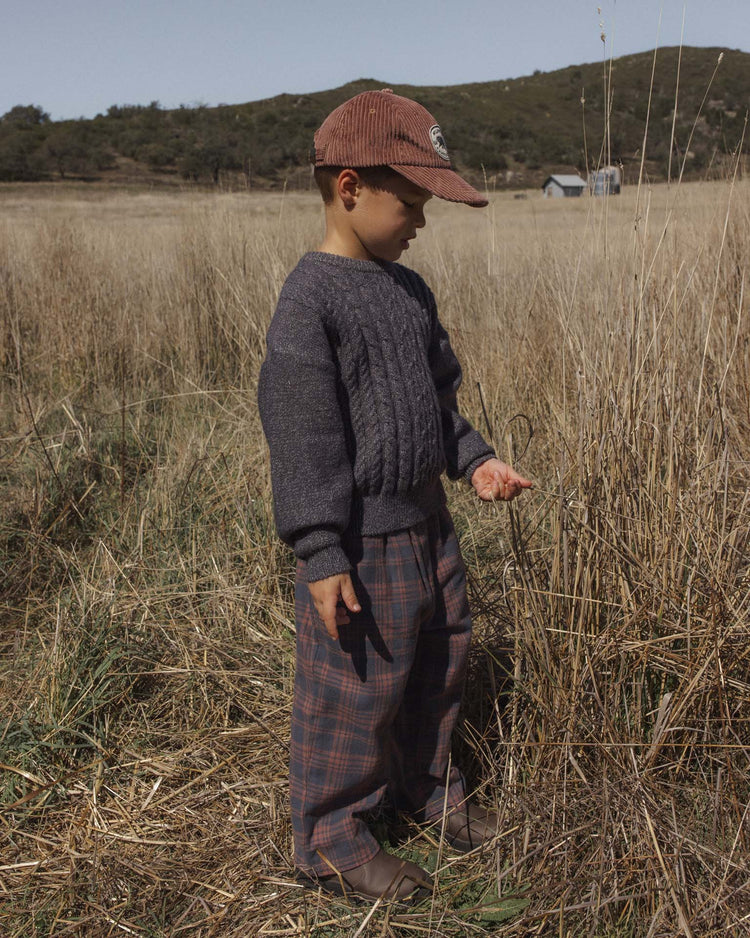A young child wearing a brown cap, gray sweater, and the rylee + cru palmer pant in midnight plaid with a baggy fit stands in a dry grassy field, looking at something in their hand, with hills and a small building in the background.