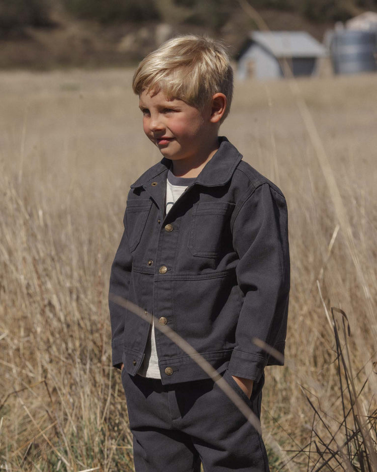 A young child with blond hair stands in a field of tall, dry grass, wearing the rylee + cru chore coat in midnight with matching pants. A few small buildings and trees appear in the softly blurred background.