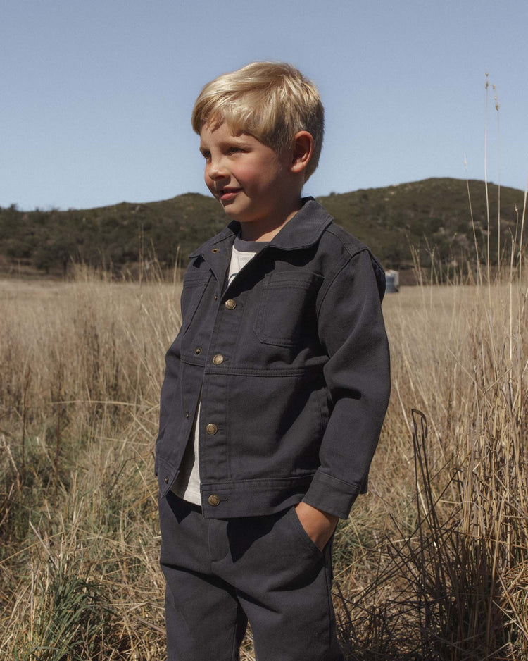 A young boy with blond hair stands in a grassy field, wearing the rylee + cru chore coat in midnight and matching pants, his hands tucked into his pockets. Rolling hills and a clear sky can be seen in the background.