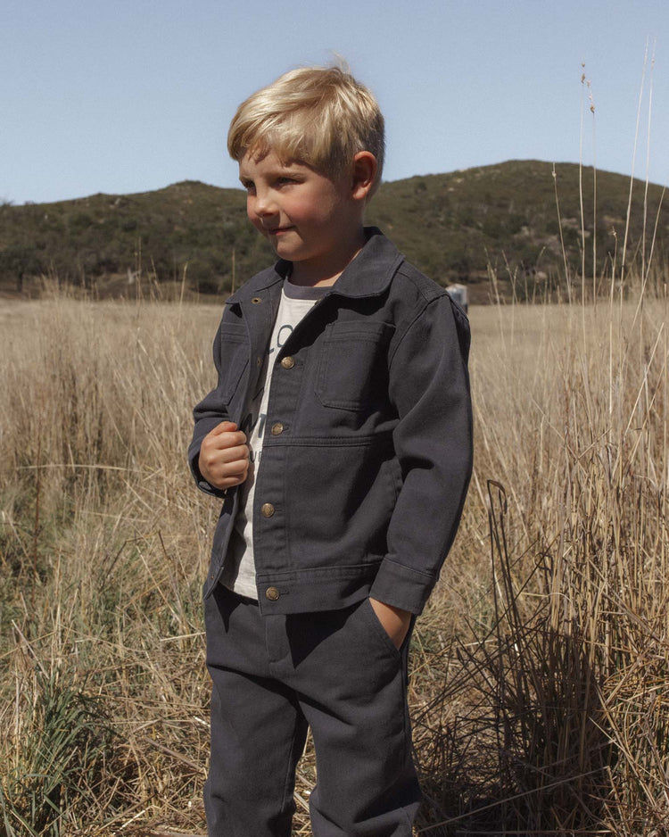 A young boy with blond hair wears the rylee + cru chore coat in midnight and matching pants, standing in a dry grassy field with hills and a clear blue sky in the background.