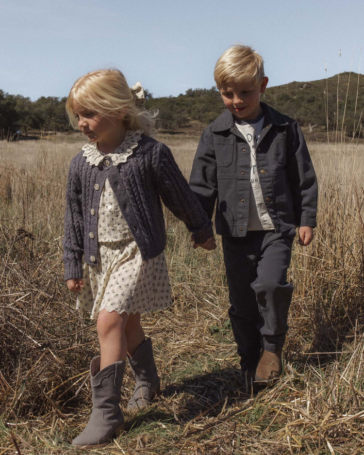 Two young children, a girl in a dress and the rylee + cru cheyenne cardigan in midnight with marble buttons and boots, and a boy in a jacket and pants, walk hand in hand through a dry grassy field on a sunny day.