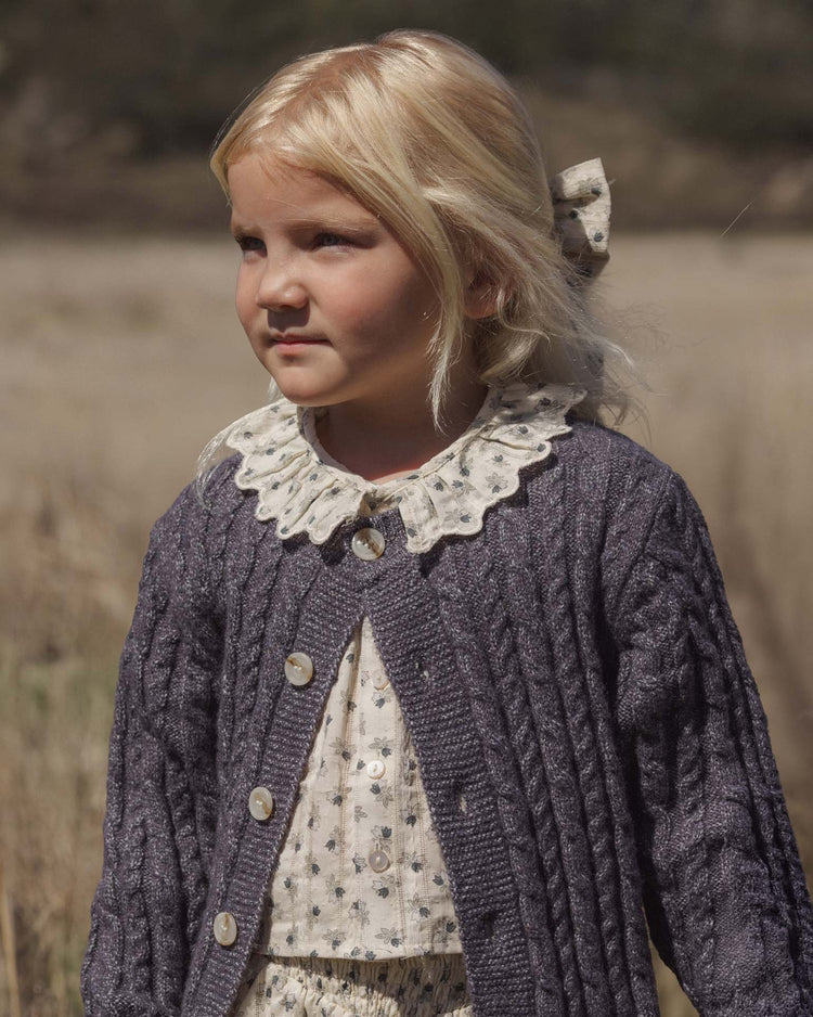 A young girl with blonde hair, wearing a floral dress with a lace collar and the rylee + cru cheyenne cardigan in midnight—adorned with marble buttons—stands outdoors in a field on a sunny day, enjoying the cozy layer.