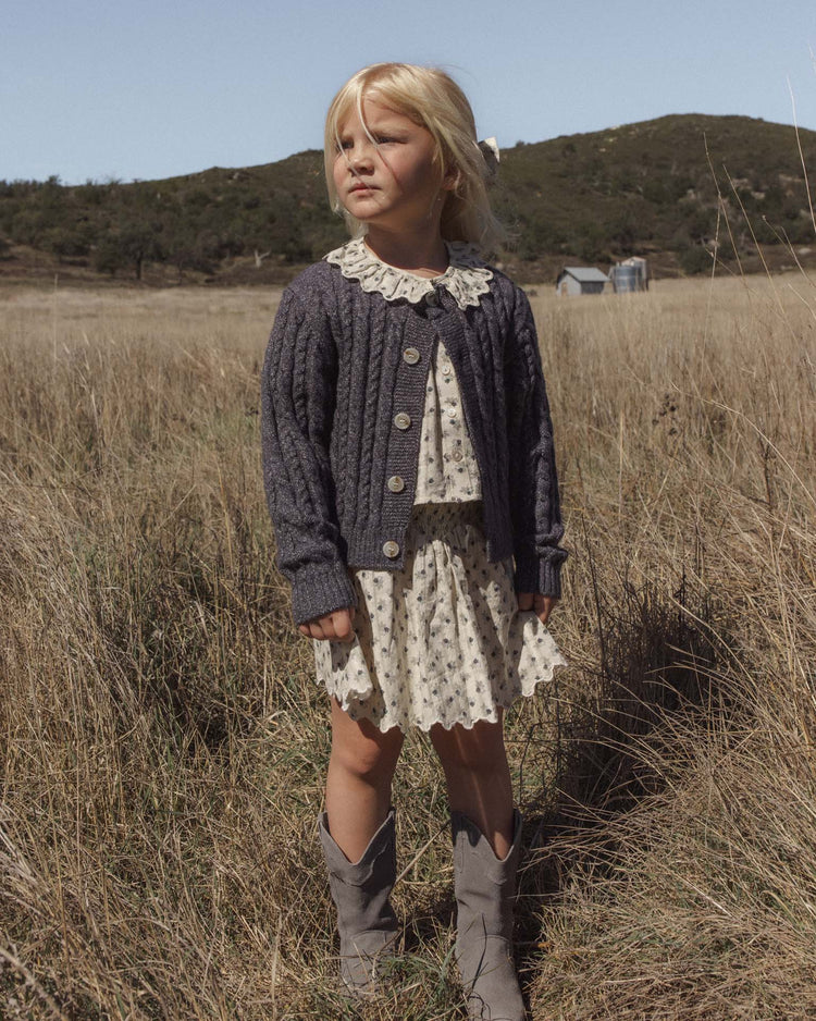 A young girl stands in a grassy field, wearing a patterned dress layered with the rylee + cru cheyenne cardigan in midnight, featuring marble buttons for a cozy touch, and paired with gray cowboy boots. Behind her, hills and buildings stretch under a clear sky.