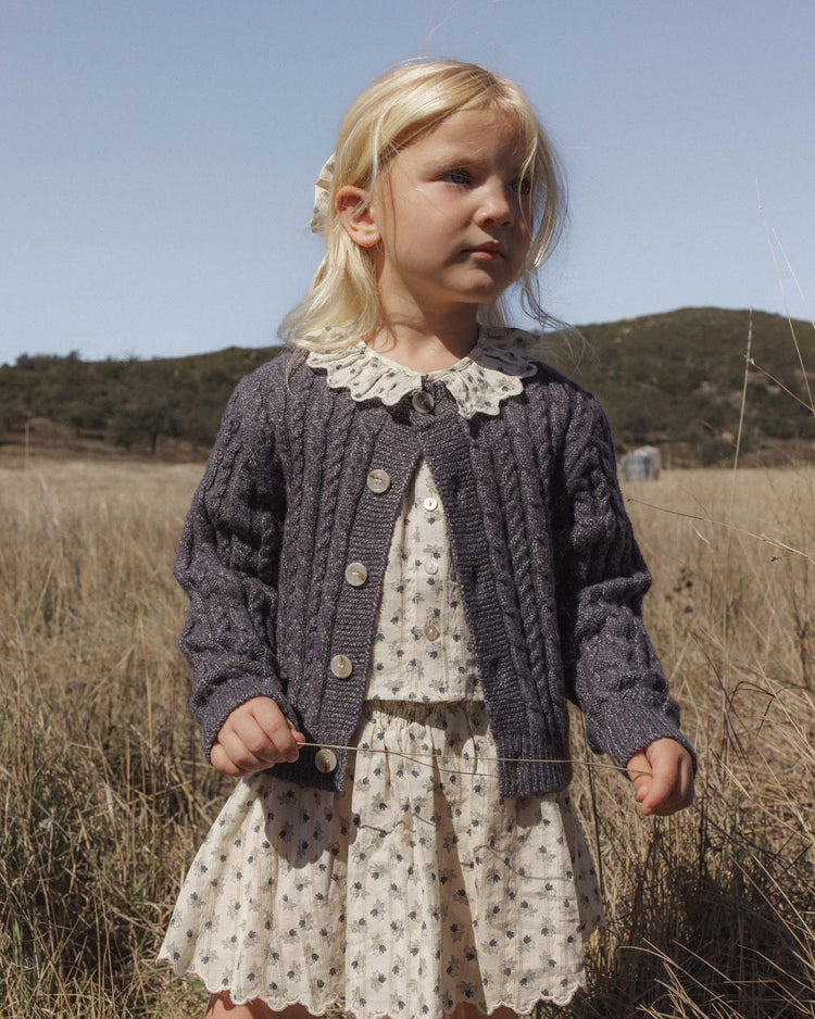 A young child with blonde hair stands in a grassy field, wearing a patterned dress with a lace collar and the rylee + cru cheyenne cardigan in midnight, fastened with delicate marble buttons. Hills and clear sky are visible in the background.
