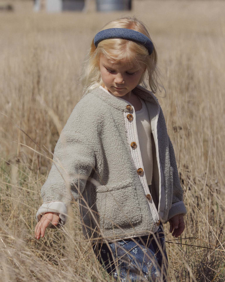 A young child with blonde hair wears the rylee + cru padded headband in indigo, standing thoughtfully in a dry grassy field while dressed in a light-colored fleece jacket and touching the tall grass.