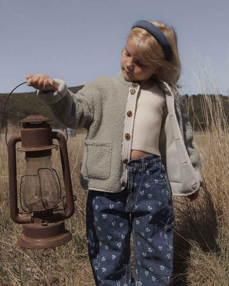 A young child wearing rylee + cru’s barrel pant in heart ditsy pairs their cozy jacket with a rustic lantern while standing in a grassy field under a clear sky.