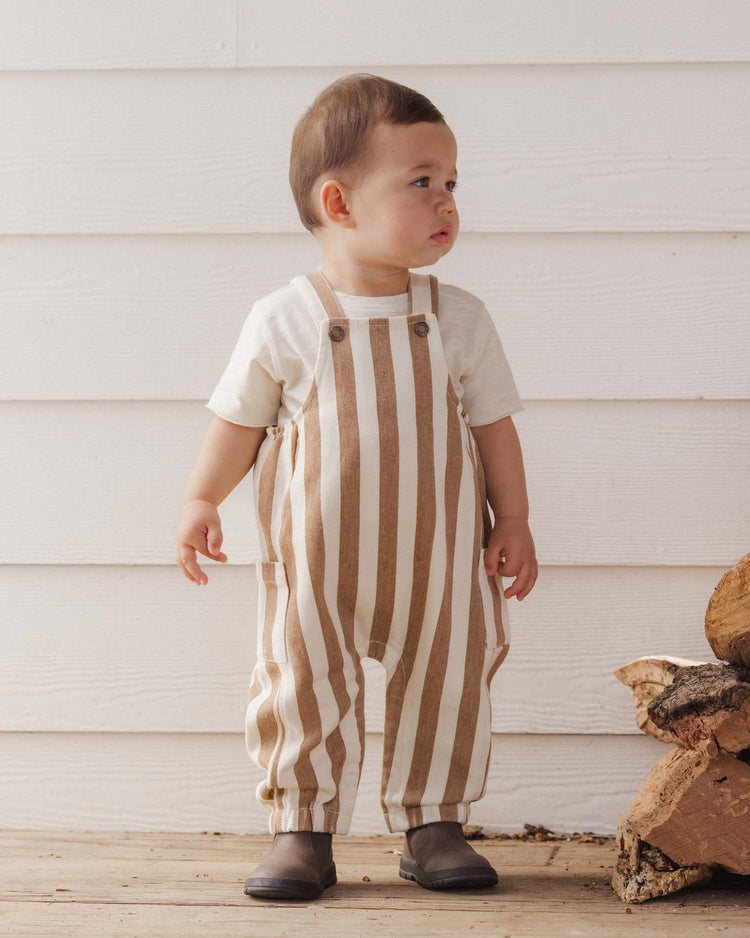 A toddler stands indoors wearing a cream shirt and the rylee + cru cargo baby overall in fawn pinstripe, crafted from 100% cotton, paired with brown boots. The child looks to the side near a stack of firewood. The background features white horizontal paneling on the wall.