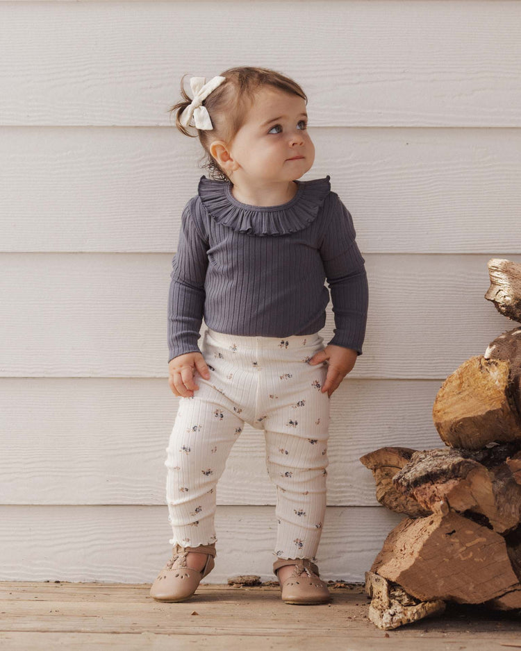 A toddler stands beside a stack of firewood, wearing the rylee + cru PREORDER ruffle collar bodysuit in midnight, paired with white patterned pants, tan shoes, and a white bow in their hair, gazing slightly upward against a white wooden wall.