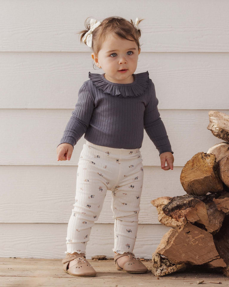 A toddler with brown hair in pigtails and white bows stands indoors by a stack of firewood, wearing the rylee + cru PREORDER ruffle collar bodysuit in midnight, cream floral leggings, and tan shoes.