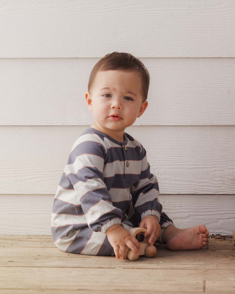 A baby with short brown hair, dressed in the rylee + cru henley jumpsuit in sage stripe made from 100% cotton, sits barefoot on a wooden floor holding a wooden toy in front of a white paneled wall.