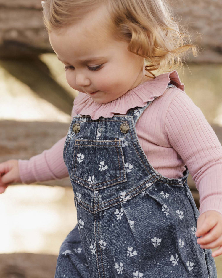 A young child with curly blonde hair wears a pink long-sleeve shirt and the rylee + cru vintage overall in heart ditsy indigo denim, looking down with a gentle expression.