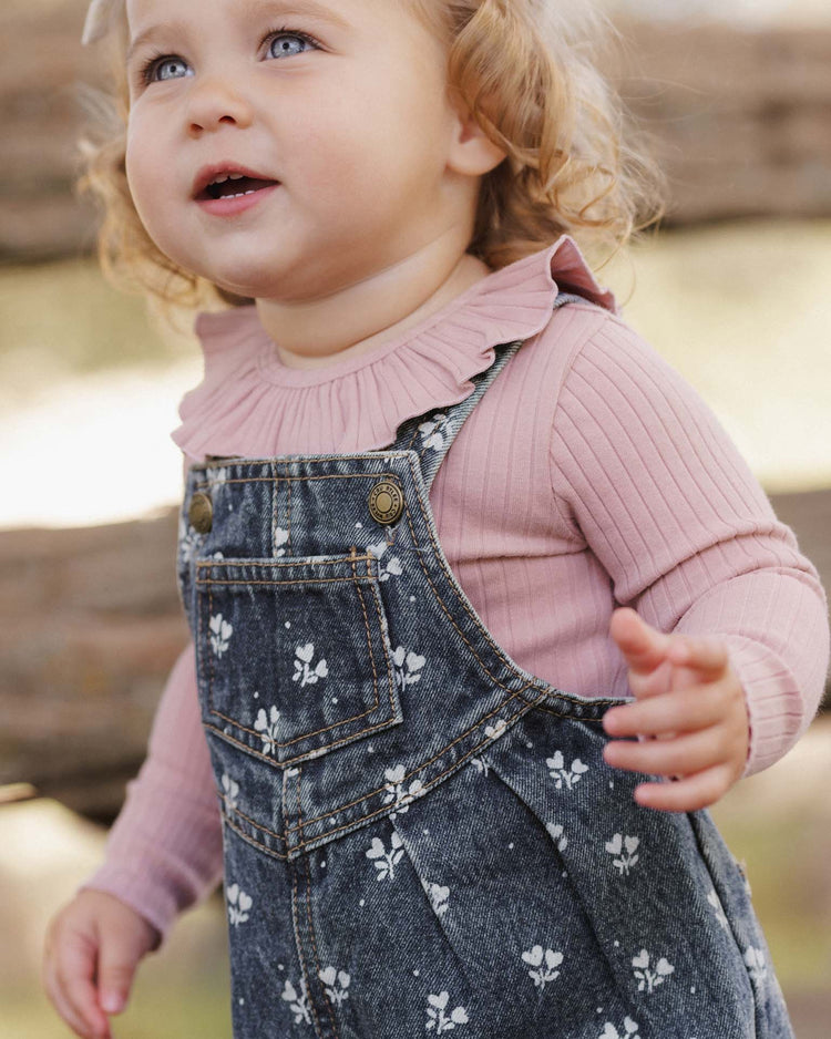 A toddler with curly blonde hair, dressed in a pink long-sleeve shirt and rylee + cru’s vintage overall in heart ditsy, crafted from 100% cotton, looks up and smiles while standing outdoors near wooden logs.