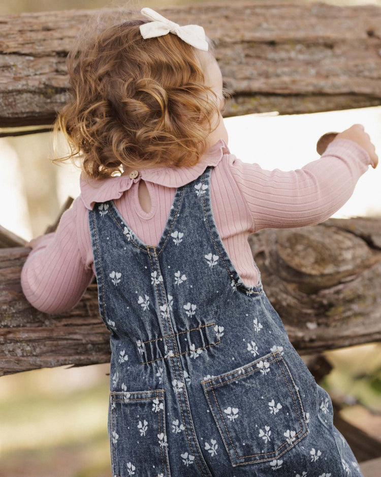 A young child with curly hair and a white bow, dressed in a pink shirt and the rylee + cru vintage overall in heart ditsy, stands facing a rustic wooden fence outdoors. Made from 100% cotton, these floral denim overalls combine comfort and timeless style for any little adventurer.