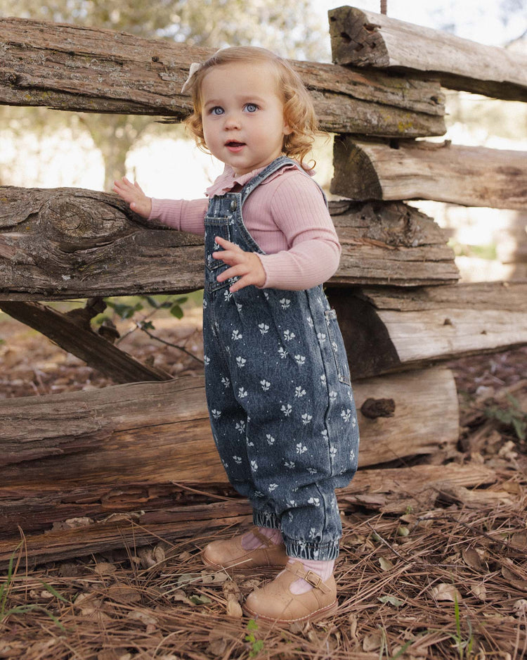 A toddler with curly blonde hair stands by a rustic wooden fence outdoors, wearing a pink top and the rylee + cru vintage overall in heart ditsy, made from 100% cotton.