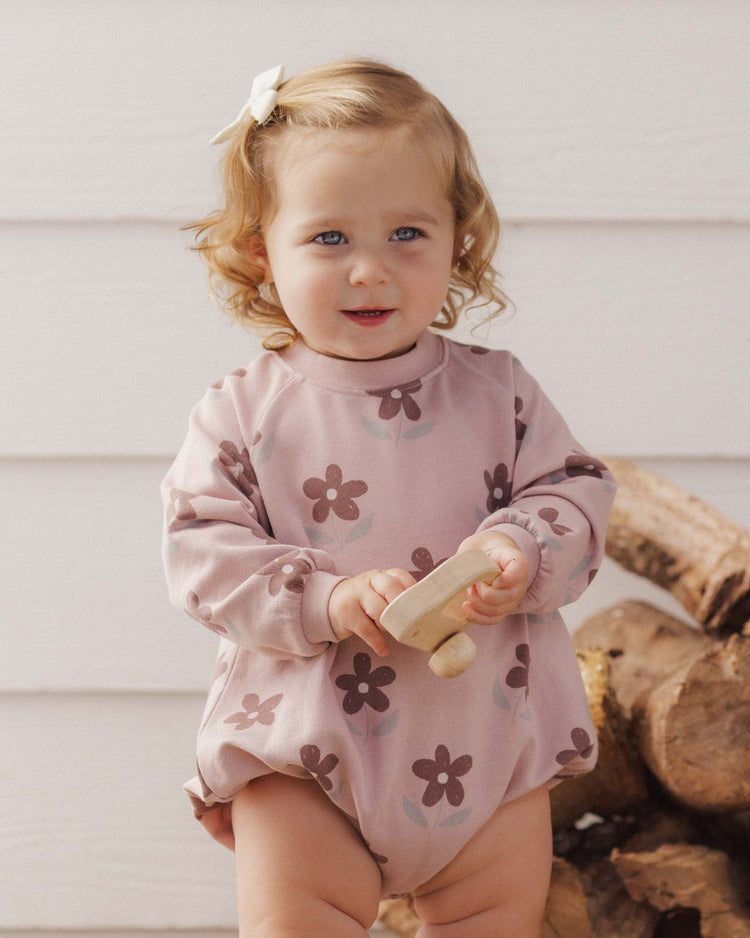 A toddler with curly blonde hair, dressed in the rylee + cru PREORDER crew neck romper in daisy and a matching hair bow, stands in front of stacked firewood while holding a wooden toy.