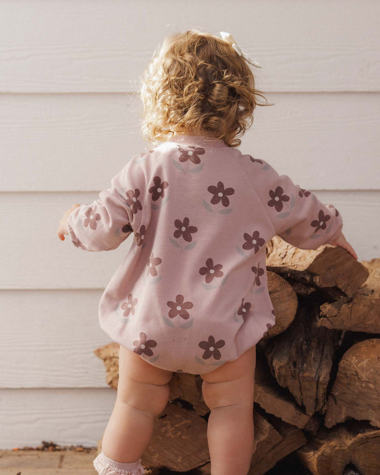 A toddler with curly blonde hair, wearing the rylee + cru PREORDER crew neck romper in daisy and socks, stands facing away while touching a stack of firewood against a white wooden wall.