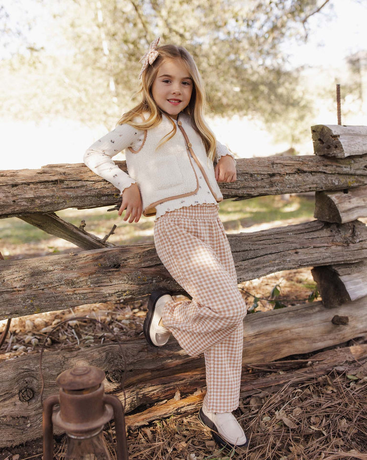 A young girl with long blonde hair leans against a rustic wooden fence outdoors, wearing the rylee + cru wide leg pant in fawn gingham with an elastic waistband, paired with a cream vest, a printed shirt, and white shoes. An old lantern sits on the ground nearby.
