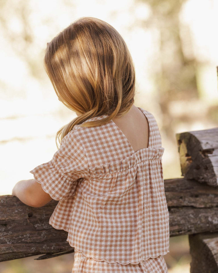 A young child with long blond hair wears the rylee + cru francie blouse in fawn gingham, standing outdoors with their back to the camera and leaning on a wooden fence as sunlight softly illuminates the natural background.