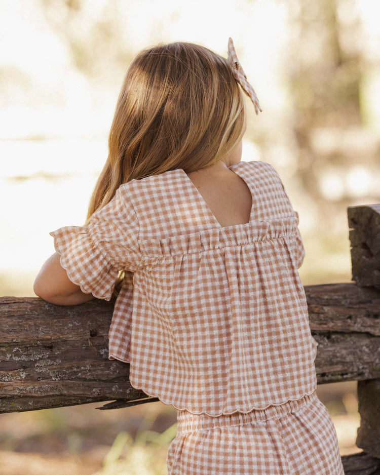 A young girl with long, light brown hair stands outdoors, leaning on a rustic wooden fence with her back to the camera. She wears the rylee + cru francie blouse in fawn gingham, styled in a cropped fit and paired with a matching bow in her hair.